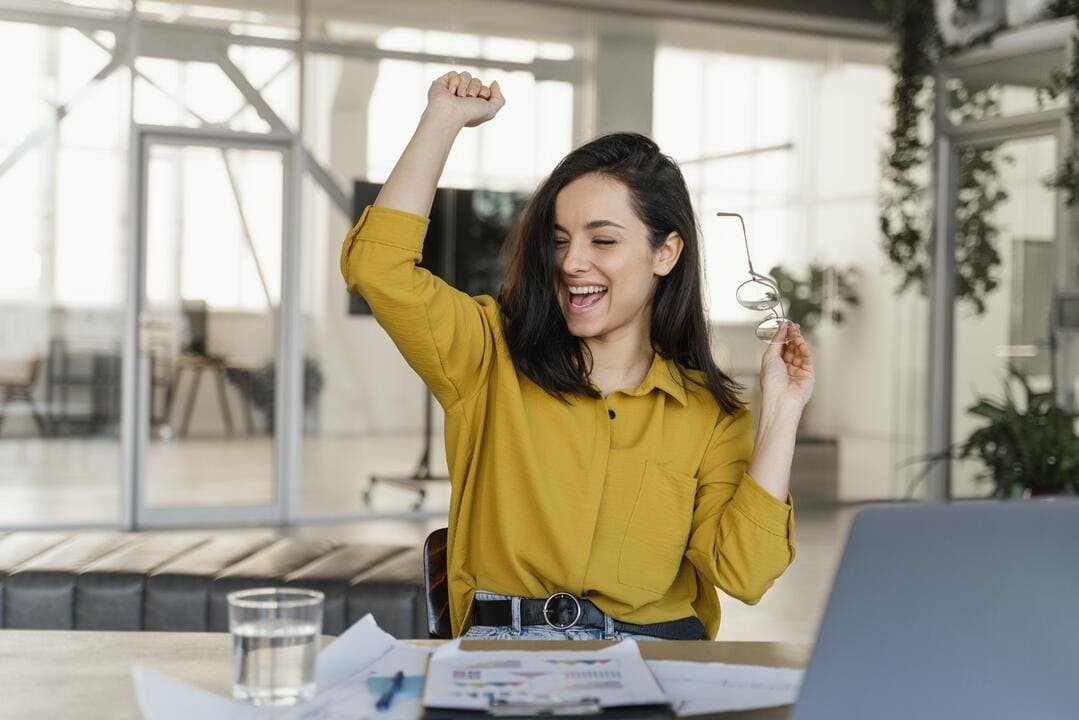 Uma mulher jovem está sentada à mesa em um ambiente moderno de escritório, com expressão de alegria e comemoração. Ela veste uma camisa amarela e segura um par de óculos com uma mão enquanto levanta o outro braço em um gesto de vitória. Sobre a mesa, há papéis espalhados e um copo de água, com um laptop parcialmente visível. O fundo do ambiente possui paredes de vidro e plantas decorativas.