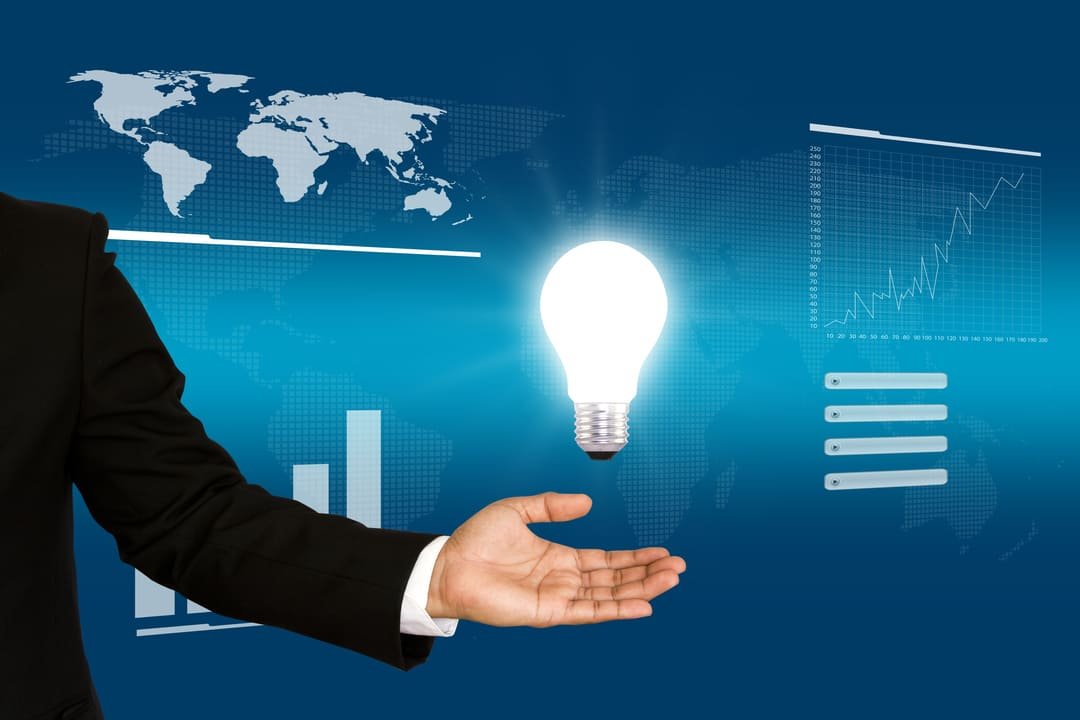 Uma mão masculina estendida, vestindo um terno preto, segura uma lâmpada brilhante que flutua acima dela. O fundo azul exibe gráficos financeiros, um mapa-múndi e indicadores de crescimento, transmitindo uma ideia de inovação e estratégia empresarial.
